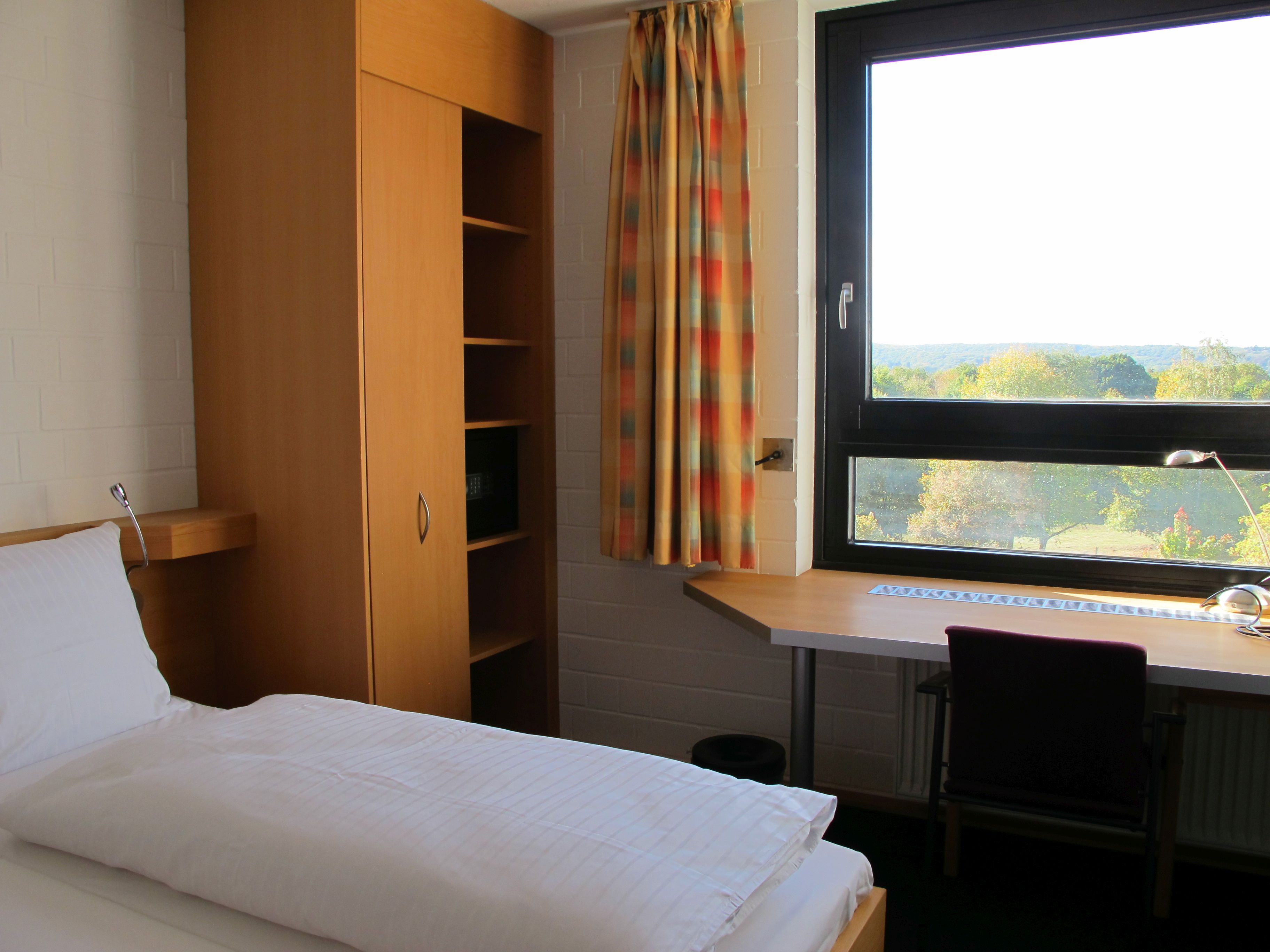 Zimmer in der FortAFin, links steht ein Bett und es ein Fenster mit Aussicht auf den Wald zu sehen, davor steht ein Schreibtisch