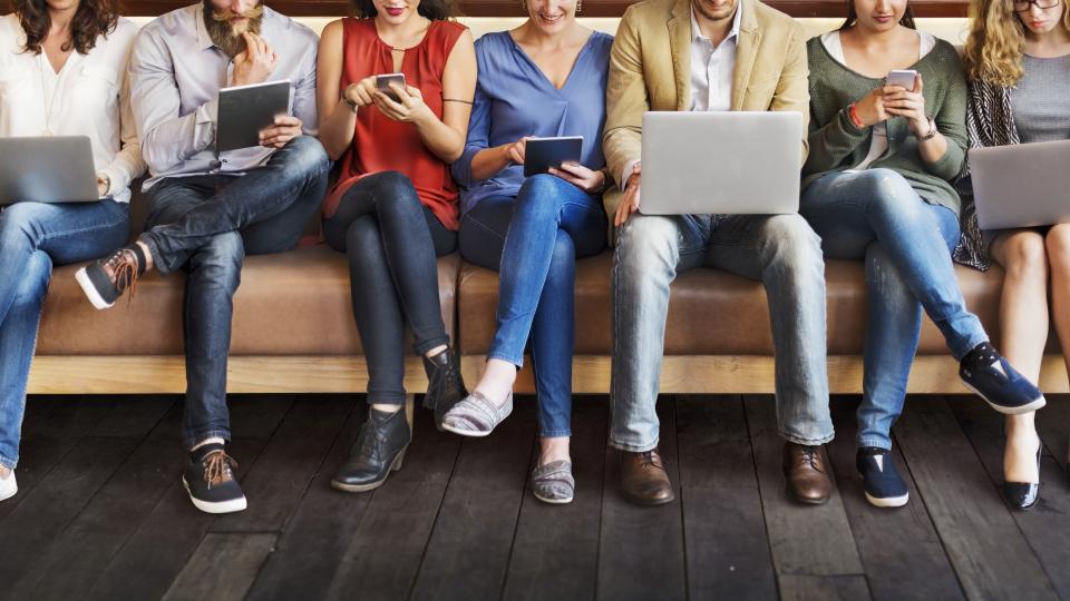 Gruppe von Menschen sitzt auf einer langen Couch mit Laptops, Tablets und Smartphones in der Hand.