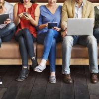 Gruppe von Menschen sitzt auf einer langen Couch mit Laptops, Tablets und Smartphones in der Hand.