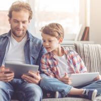 Ein Vater sitzt mit seinem Sohn auf einem Sofa, beide schauen gemeinsam auf ein Tablet.